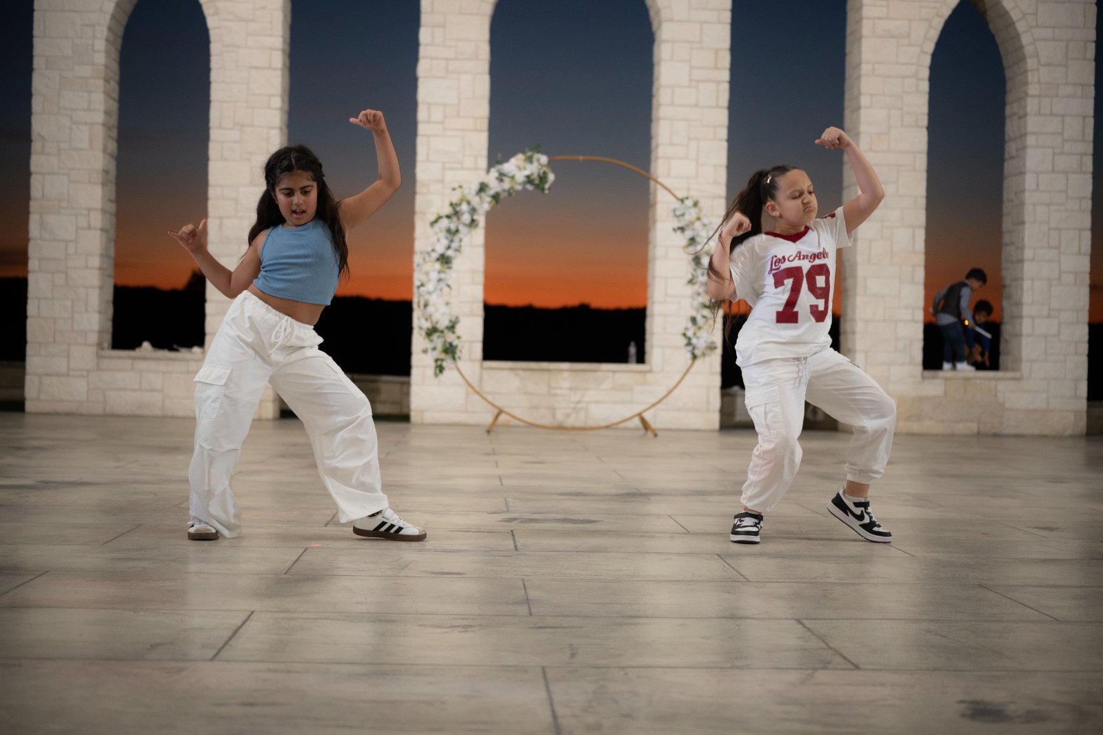 Two kids performing a dance routine outdoors at sunset in front of a floral arch, captured by Zan Visuals during an event celebration.