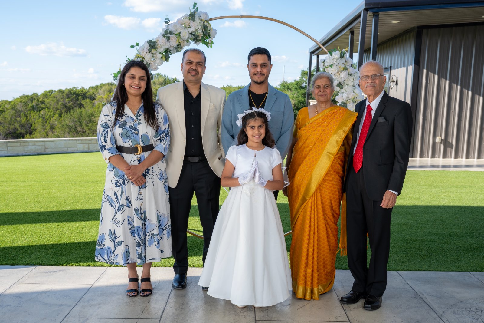 Family group photo under a white floral arch during a First Communion celebration, featuring the child in her white dress surrounded by relatives, captured by Zan Visuals as part of professional event photography.