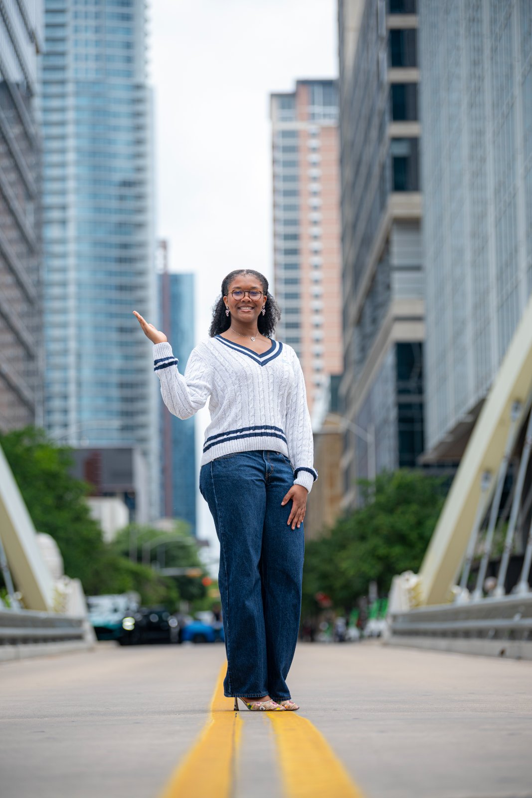 Graduate standing on a city bridge between tall buildings, wearing a white sweater and jeans, captured by Zan Visuals during an urban graduation photo session.