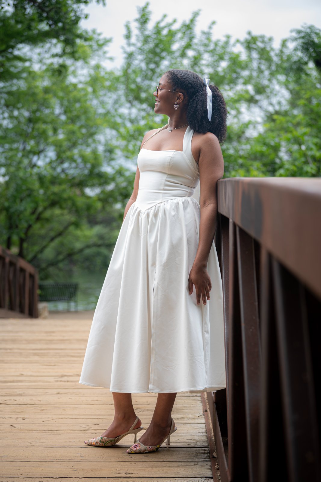 Graduate posing on a wooden bridge in a white dress surrounded by greenery, captured by Zan Visuals during an outdoor graduation photo session.
