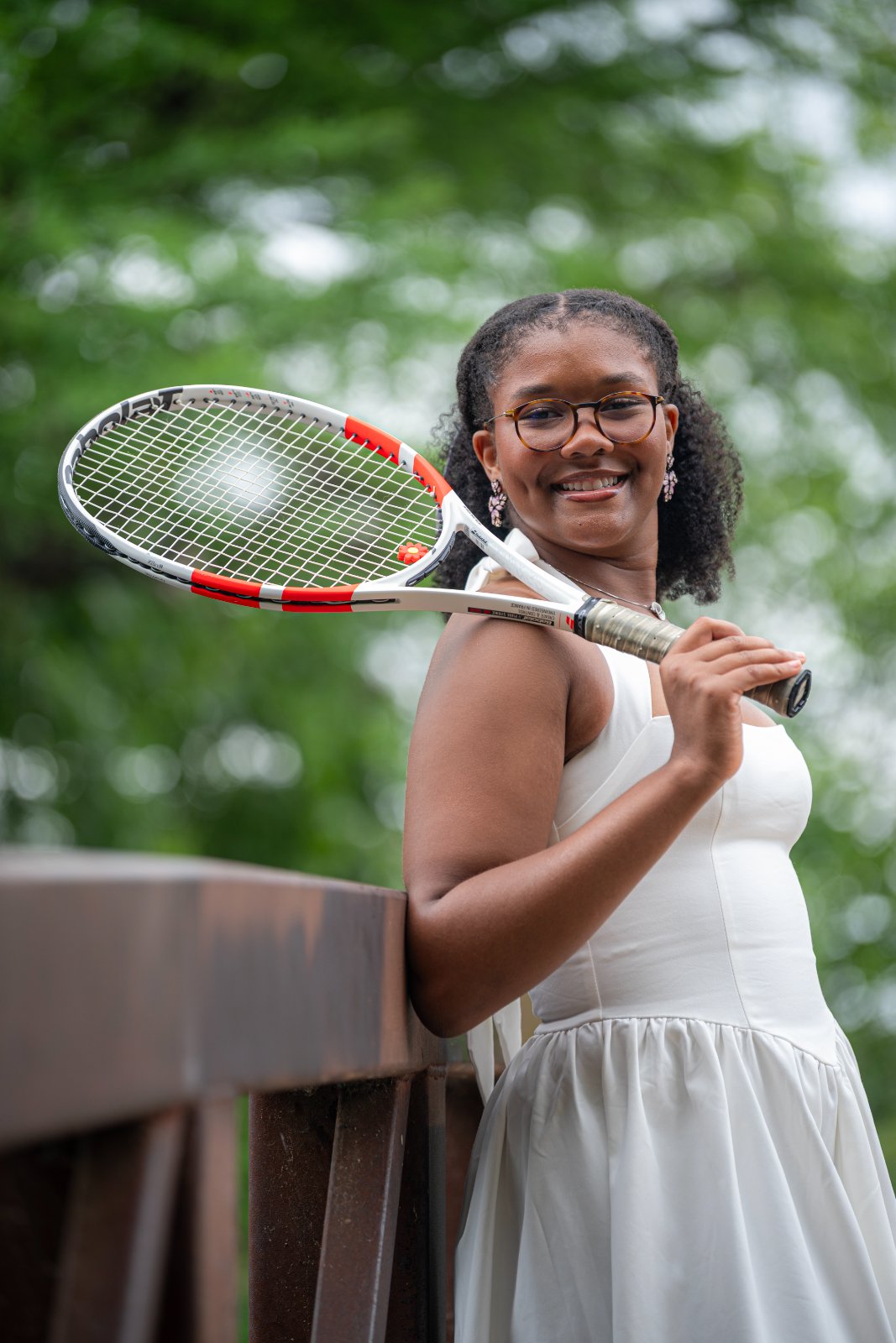 Graduate posing on a wooden bridge holding a tennis racket over the shoulder, blending athletic personality with an elegant white dress, captured by Zan Visuals during an outdoor graduation photo session.
