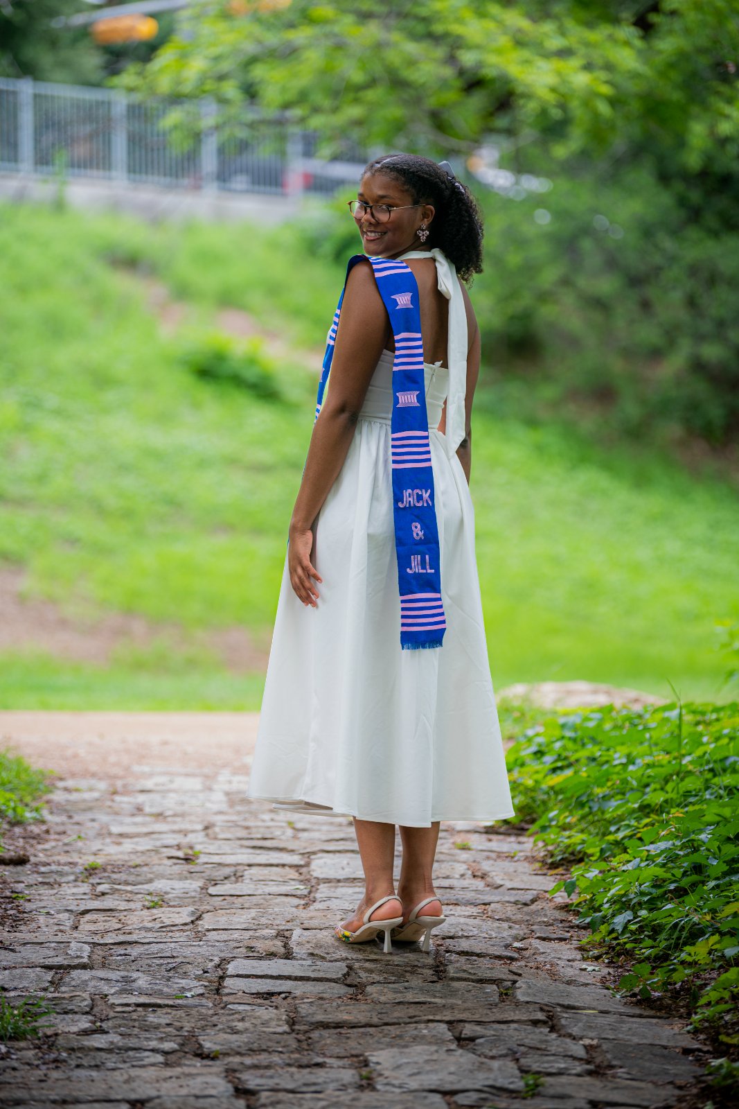 Graduate smiling over the shoulder while wearing a blue and white stole on a garden pathway, captured by Zan Visuals during an outdoor graduation photo session.