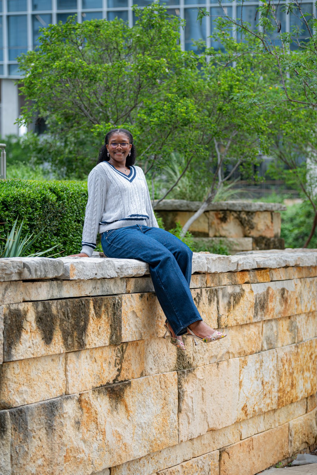 Graduate sitting on a stone wall in a landscaped outdoor area, wearing a light gray sweater and jeans, captured by Zan Visuals during a relaxed graduation photo session.