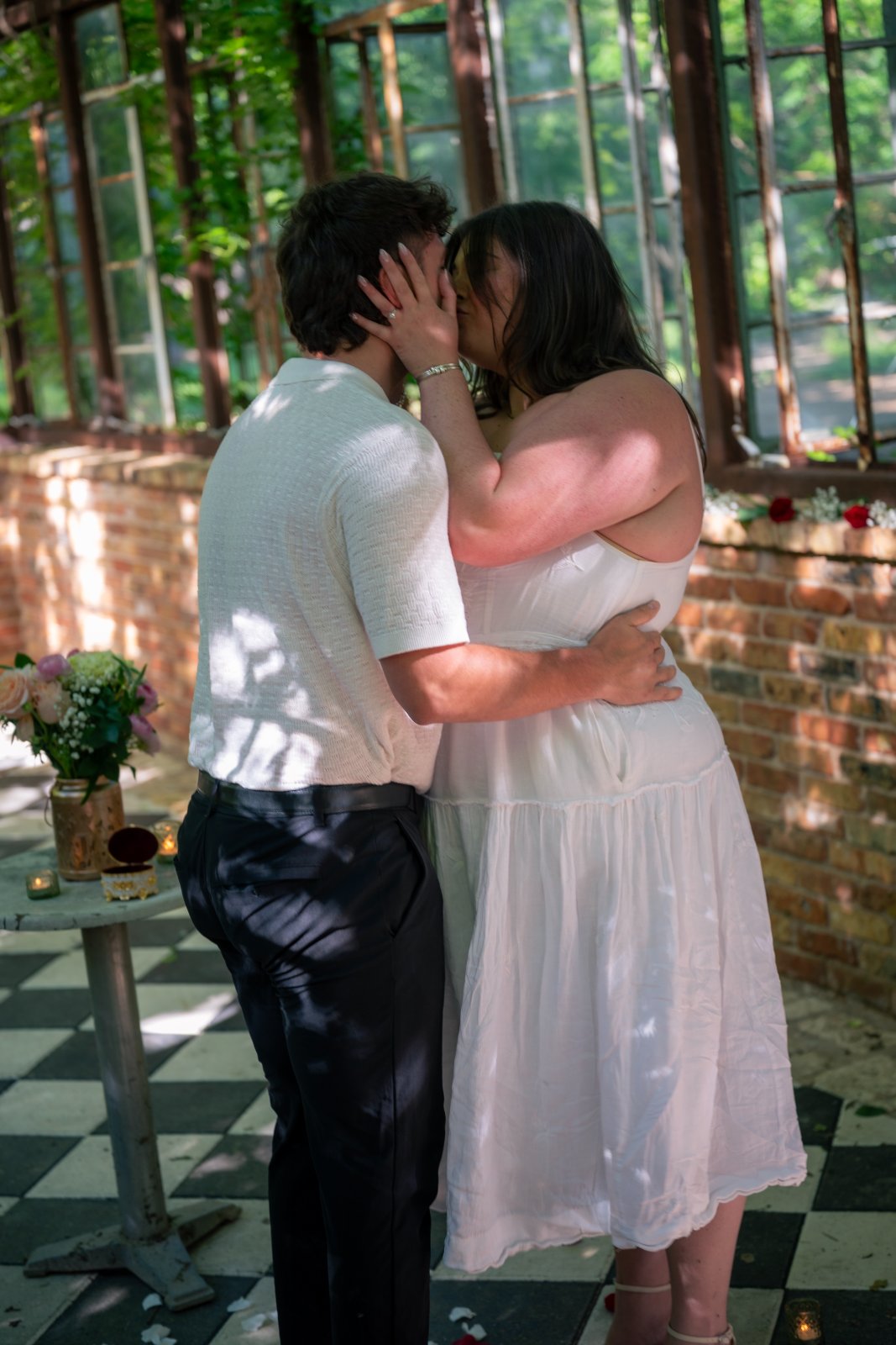 Romantic proposal moment of a couple kissing inside a sunlit greenhouse with flowers and candles — taken by Zan Visuals.