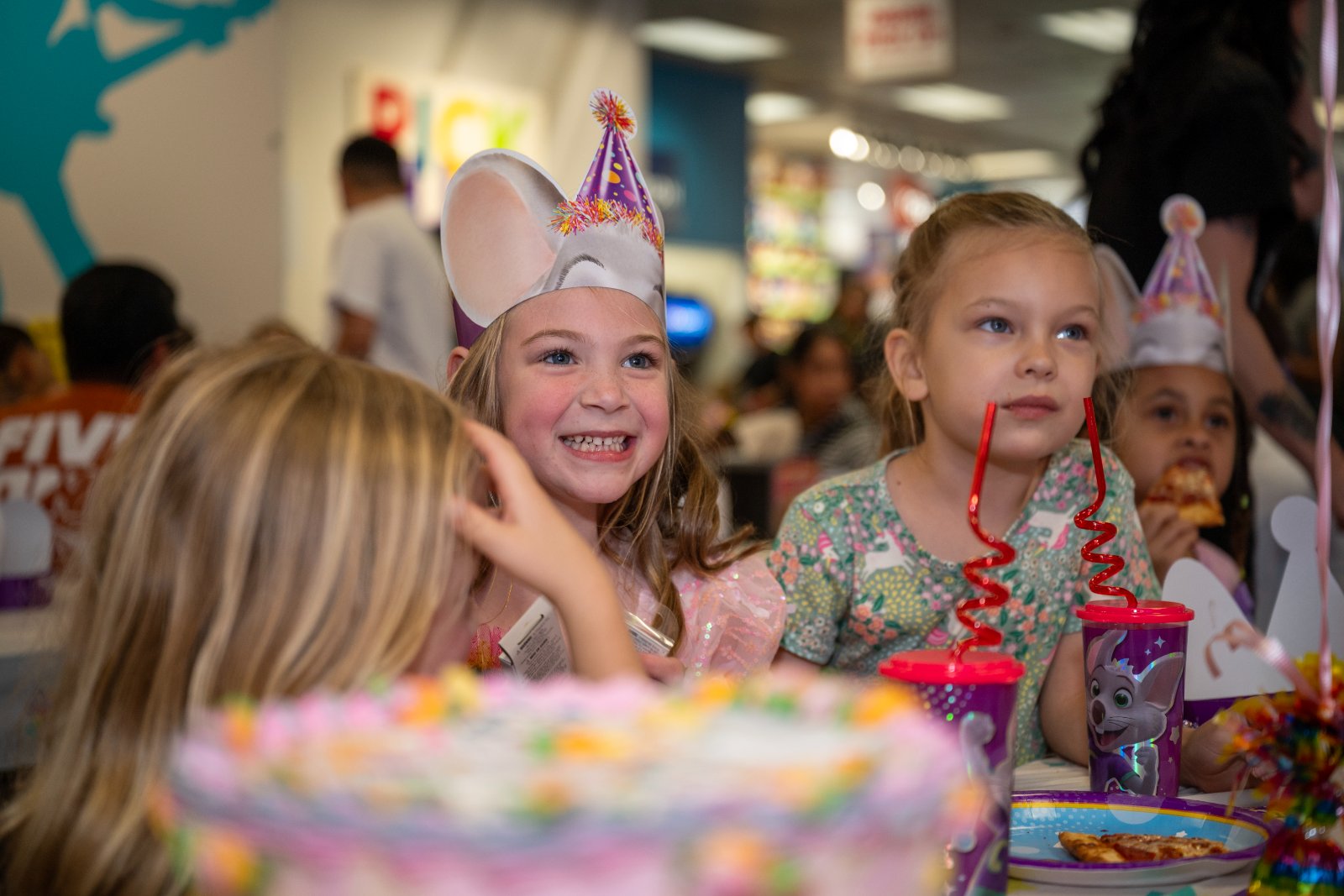 Children gathered around a party table enjoying pizza and drinks during a colorful birthday celebration, with a cake visible in the foreground — photographed by Zan Visuals.