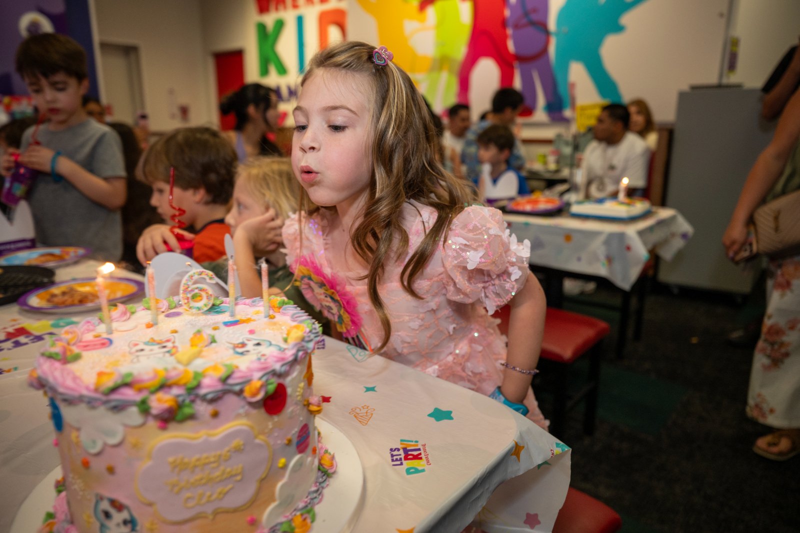 Child blowing out candles on a colorful sixth‑birthday cake during a lively kids’ party, surrounded by friends and festive decorations — photographed by Zan Visuals.