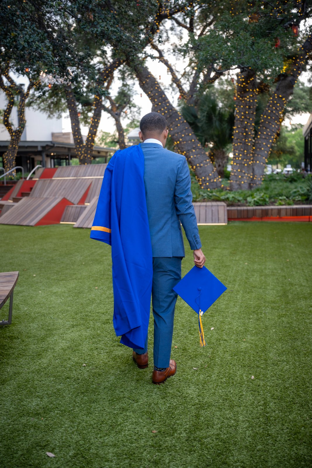 Austin TX graduation portrait showing a back‑view shot of a young man in a blue suit holding his graduation cap and gown while walking through a grassy outdoor area with trees and warm lights — taken by Zan Visuals.
