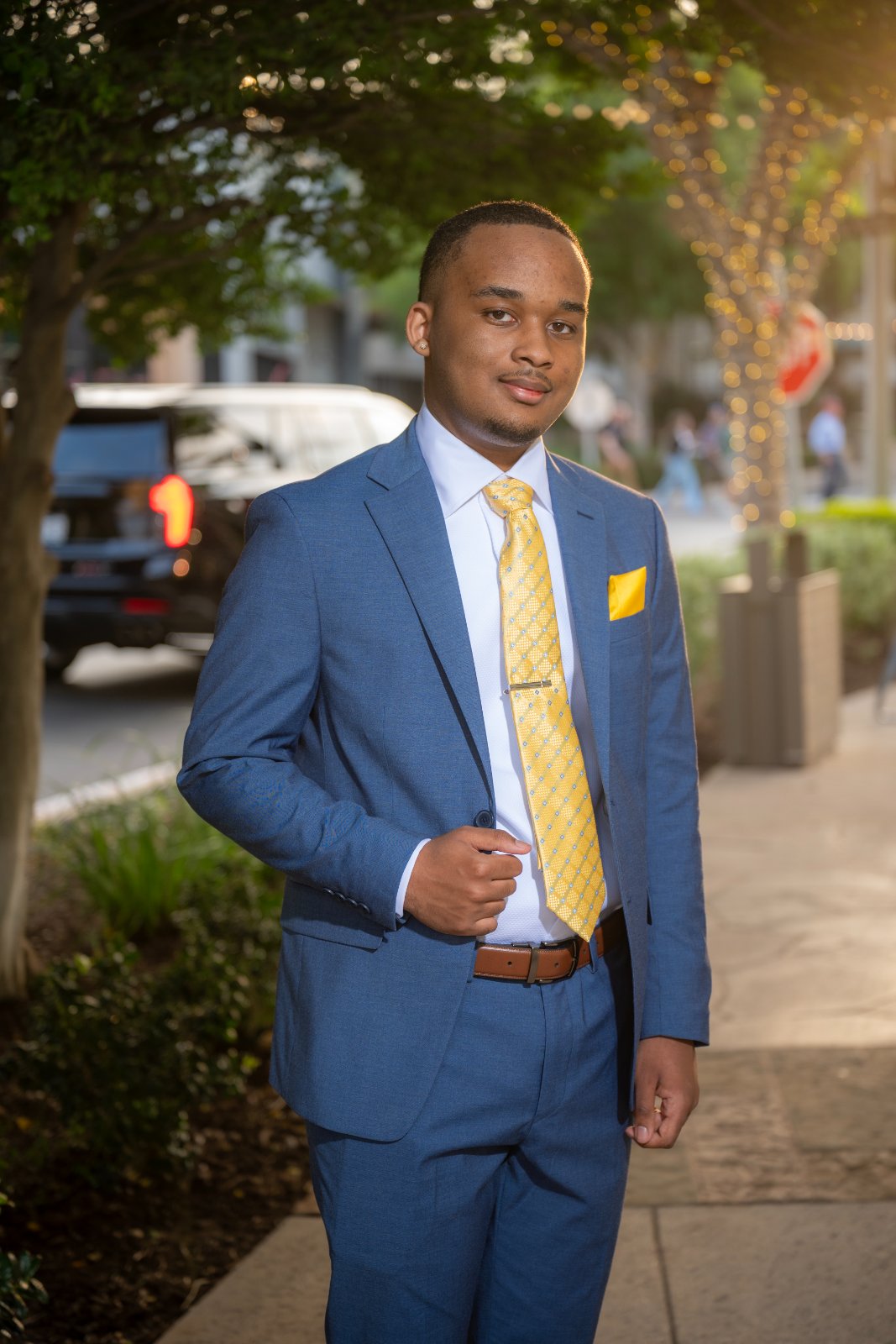 Austin TX portrait of a young man in a blue suit with a yellow tie standing on a sunlit urban sidewalk with trees and warm evening light — taken by Zan Visuals.