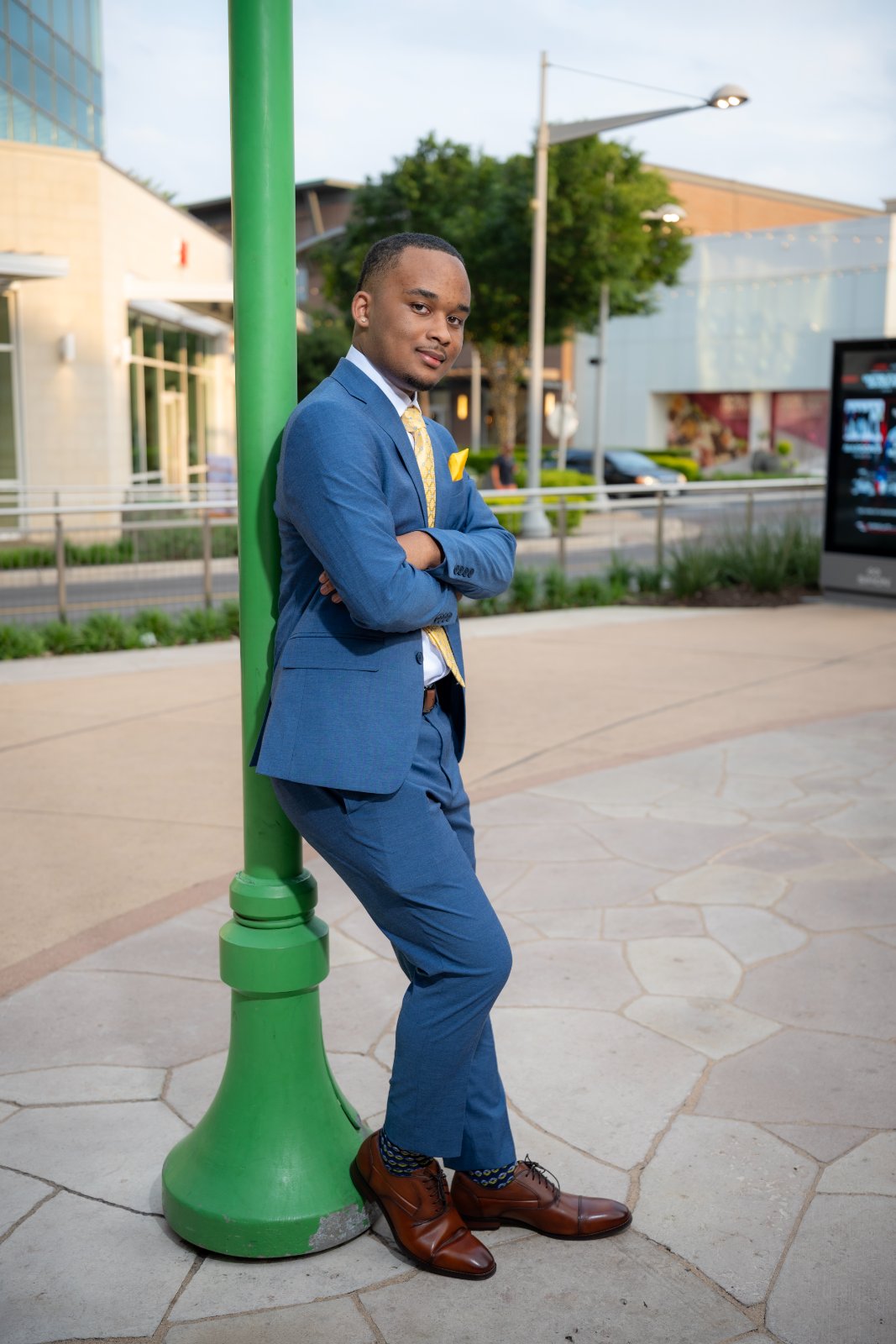 Outdoor graduation portrait in Austin TX of a young man in a blue suit leaning against a green lamppost in a modern urban setting — taken by Zan Visuals.