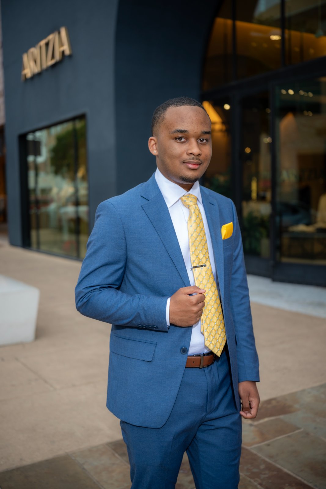 Graduation portrait in Austin TX featuring a young man in a blue suit with a yellow tie, standing confidently in front of the Aritzia storefront in natural outdoor light — taken by Zan Visuals.