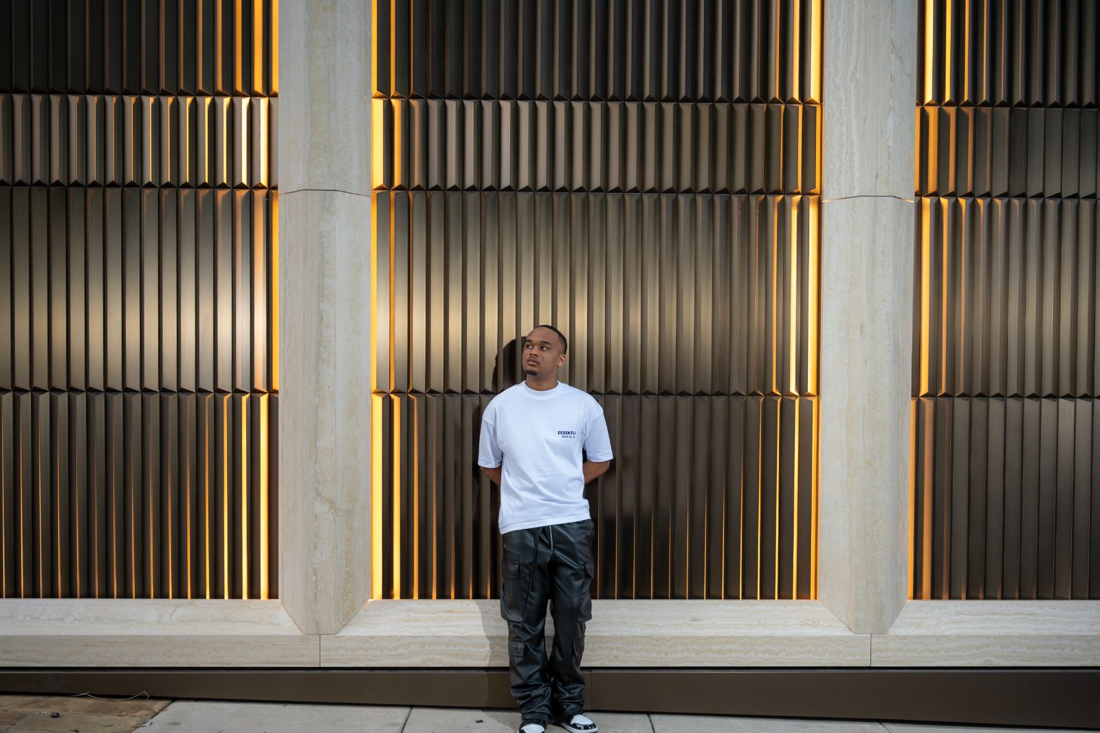 Austin TX graduation portrait of a young man wearing an Essentials shirt standing in front of a modern architectural wall with warm lighting — taken by Zan Visuals.