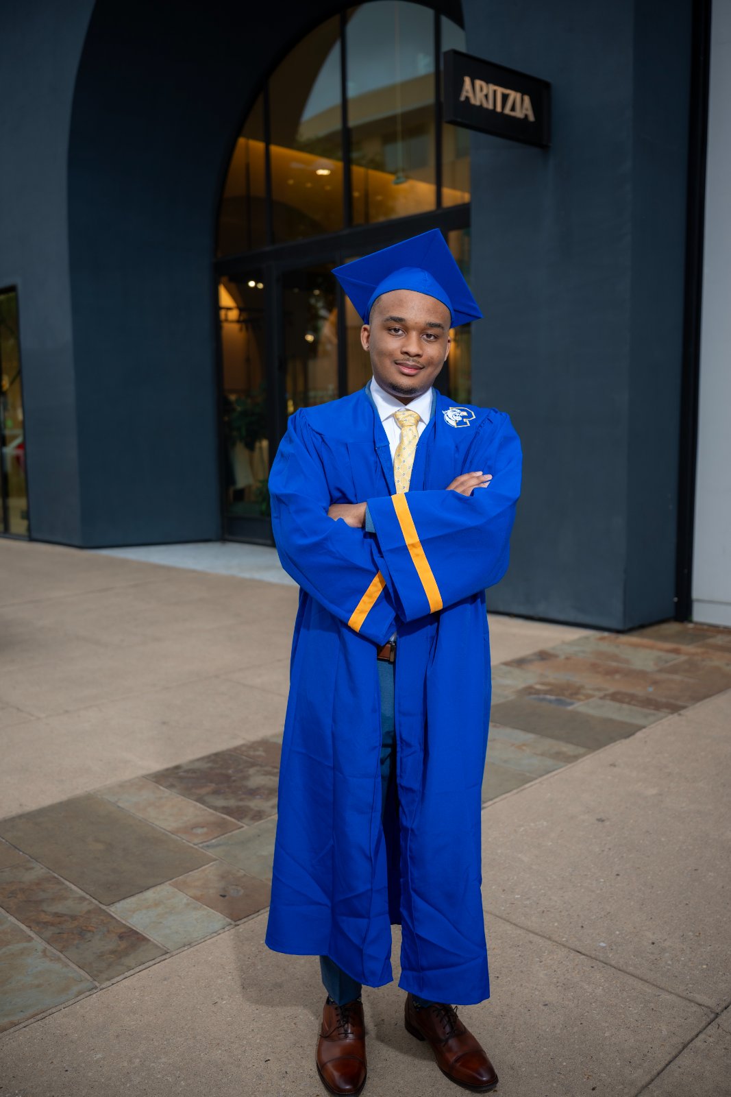 Austin TX graduation portrait of a young man in a blue cap and gown with yellow trim standing confidently in front of an arched storefront — taken by Zan Visuals.