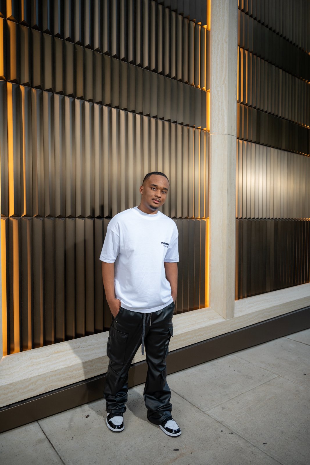 Austin TX graduation portrait of a young man wearing an Essentials shirt and black pants standing in front of a modern architectural wall with warm lighting — taken by Zan Visuals.