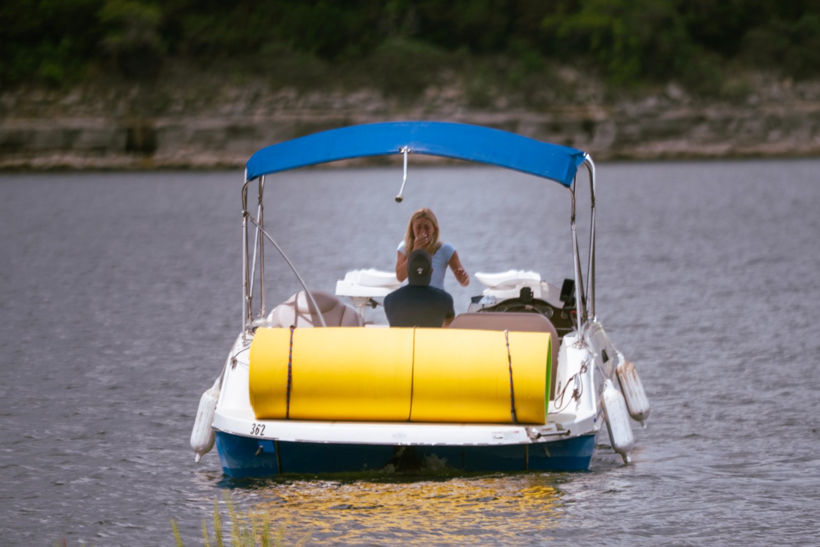 A small motorboat cruising on a calm lake with two people onboard, viewed from behind, with rocky shoreline and greenery in the background — photographed by Zan Visuals.