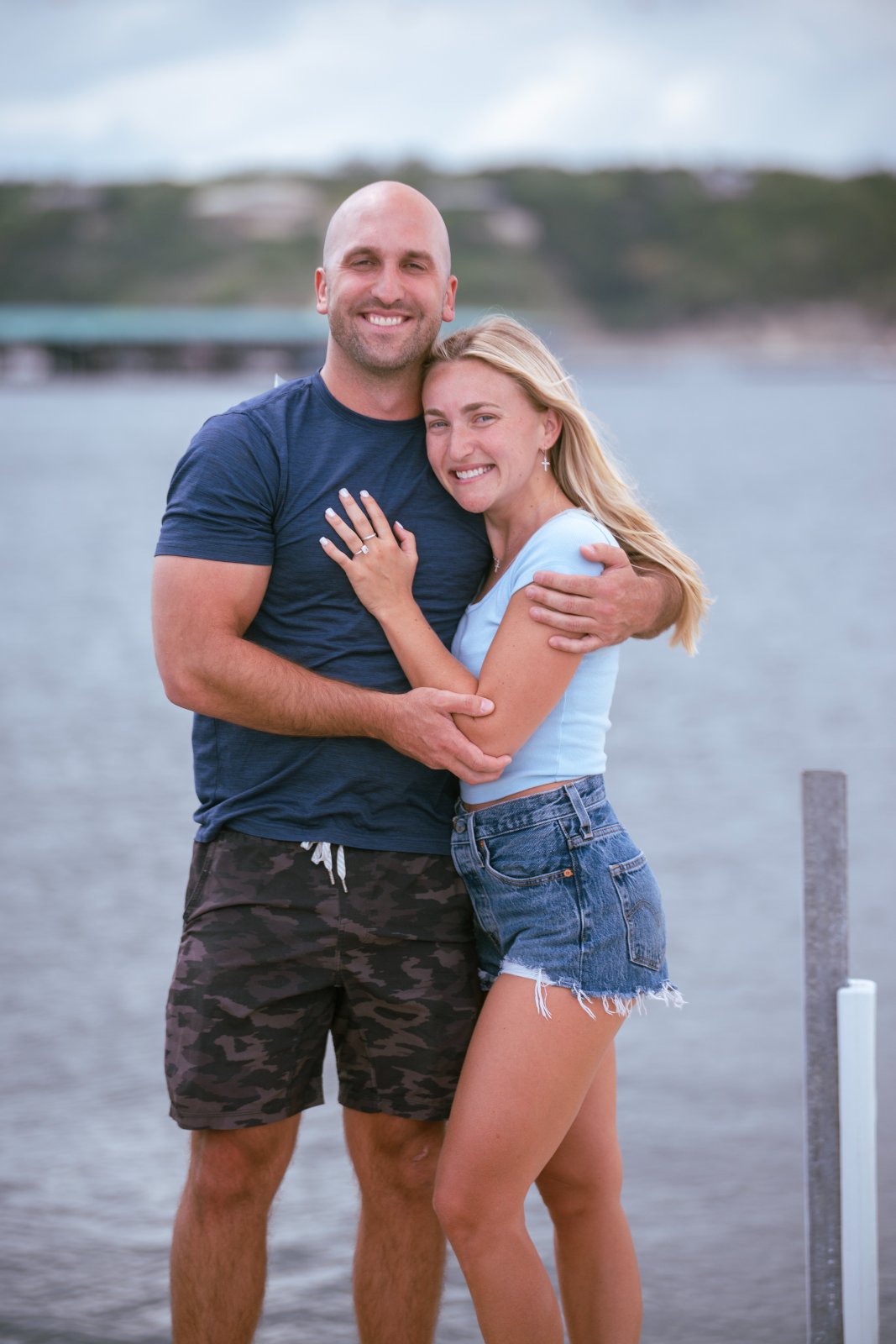 A couple embracing on a lakeside dock during a romantic proposal session, smiling together with scenic water and hills in the background — captured by Zan Visuals.