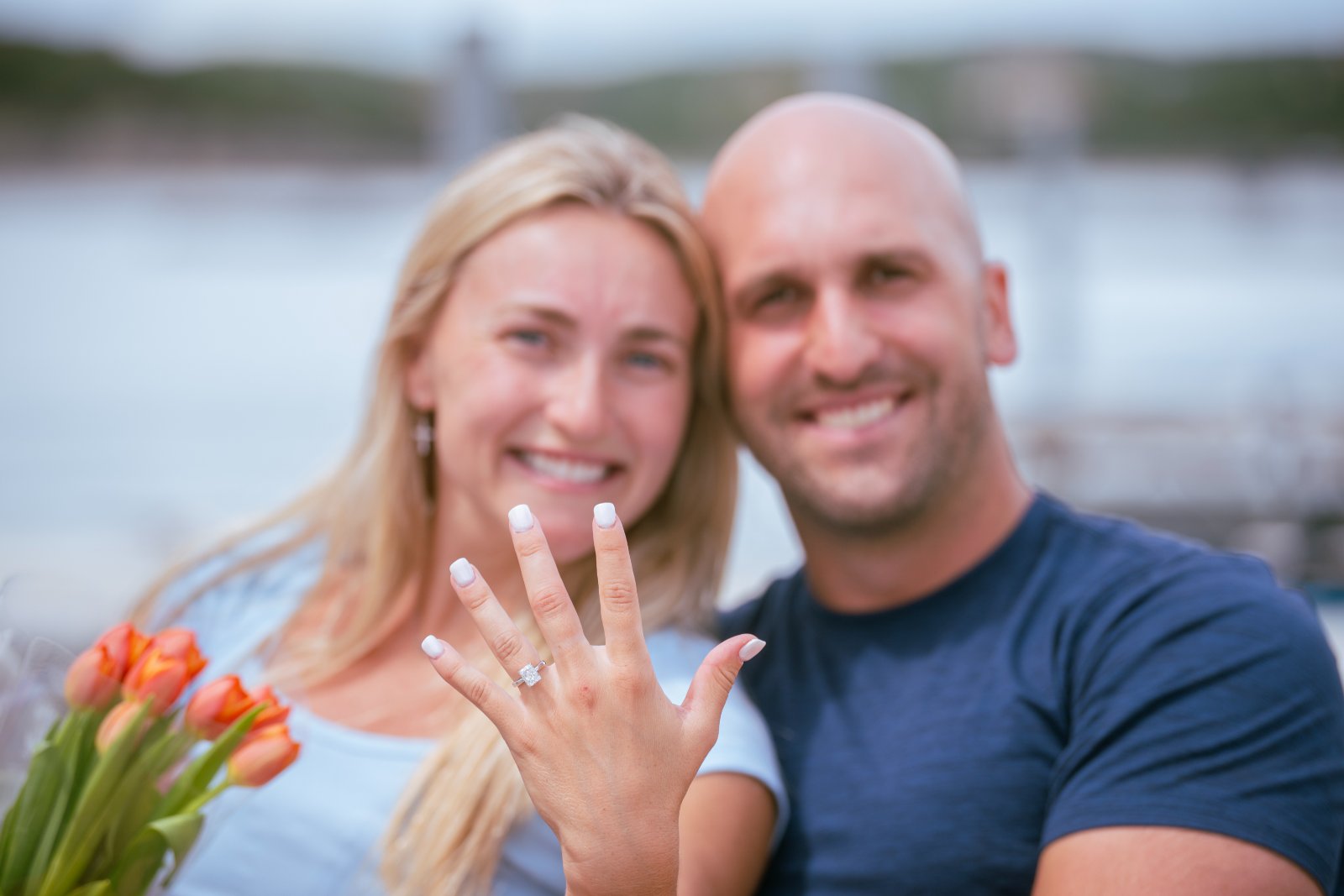A newly engaged couple sitting by the lake as the bride-to-be proudly shows her diamond ring while holding a bouquet of tulips — captured by Zan Visuals during a proposal session.