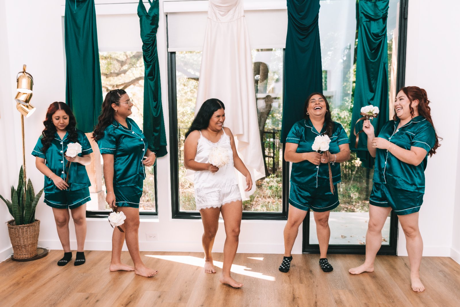 Bridesmaids in matching satin pajamas holding white bouquets, standing indoors with the bride’s dress hanging behind them, captured by Zan Visuals.