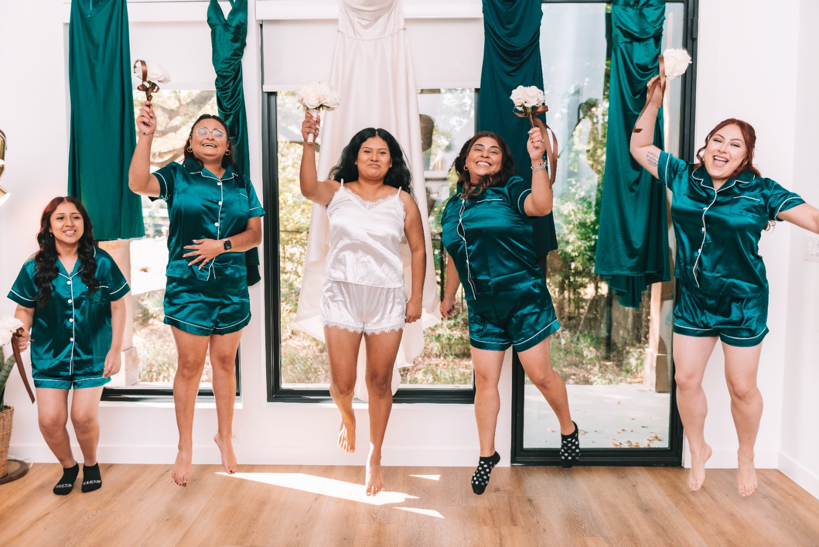 Bridesmaids in matching satin pajamas posing playfully indoors, with the bride in white holding flowers, captured by Zan Visuals.