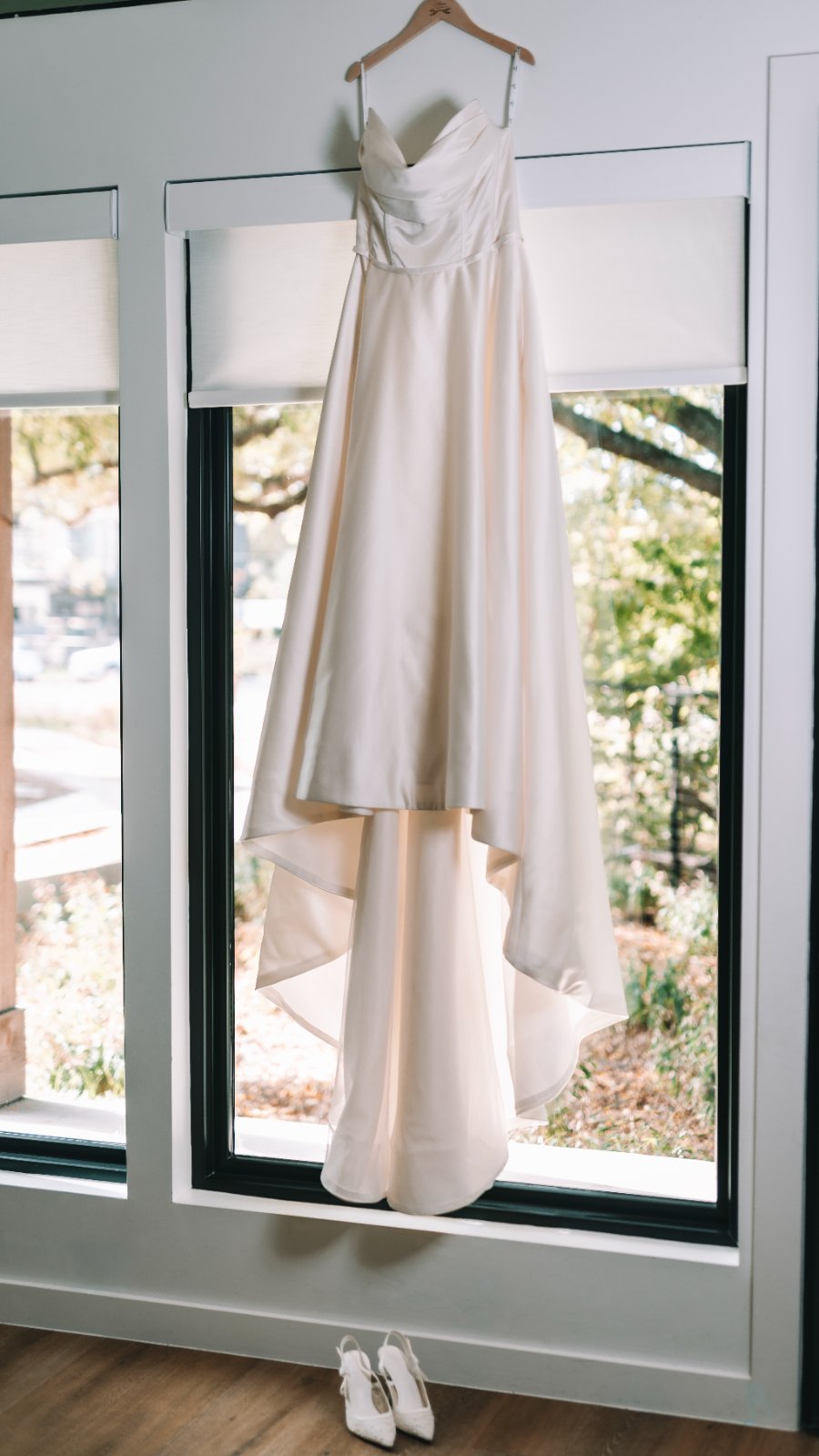 Bridal wedding dress hanging in front of a large window with matching heels placed below, captured by Zan Visuals.