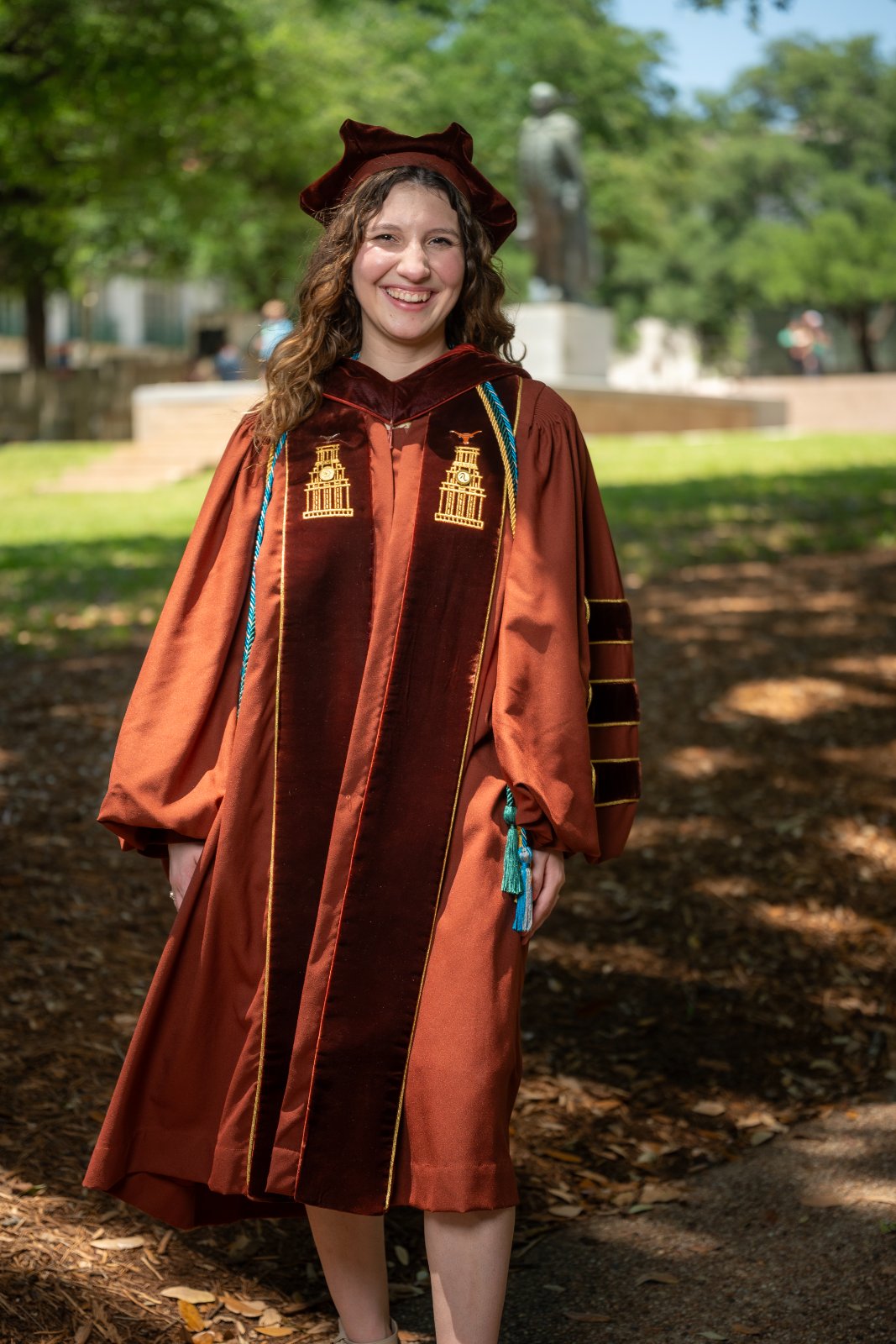 UT Austin graduation portrait of a graduate in full regalia standing along a tree‑lined campus pathway — photographed by Zan Visuals.