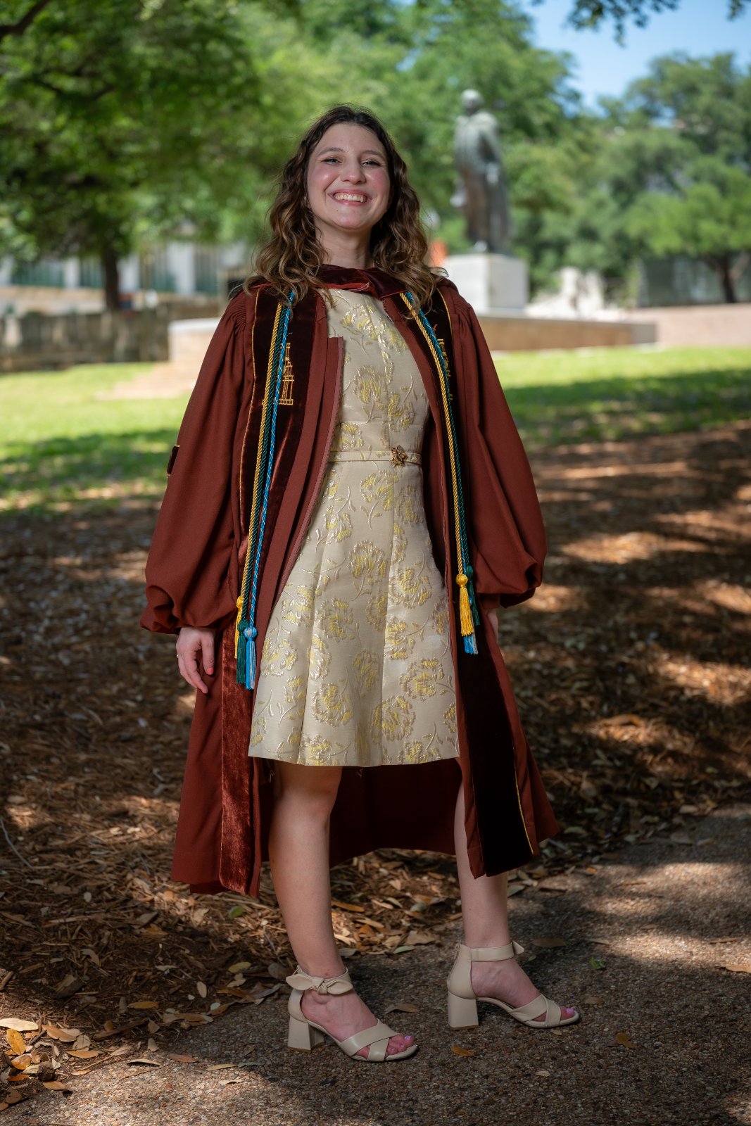 UT Austin graduation portrait featuring the graduate in a gold dress and regalia standing along a tree‑lined campus pathway — photographed by Zan Visuals.