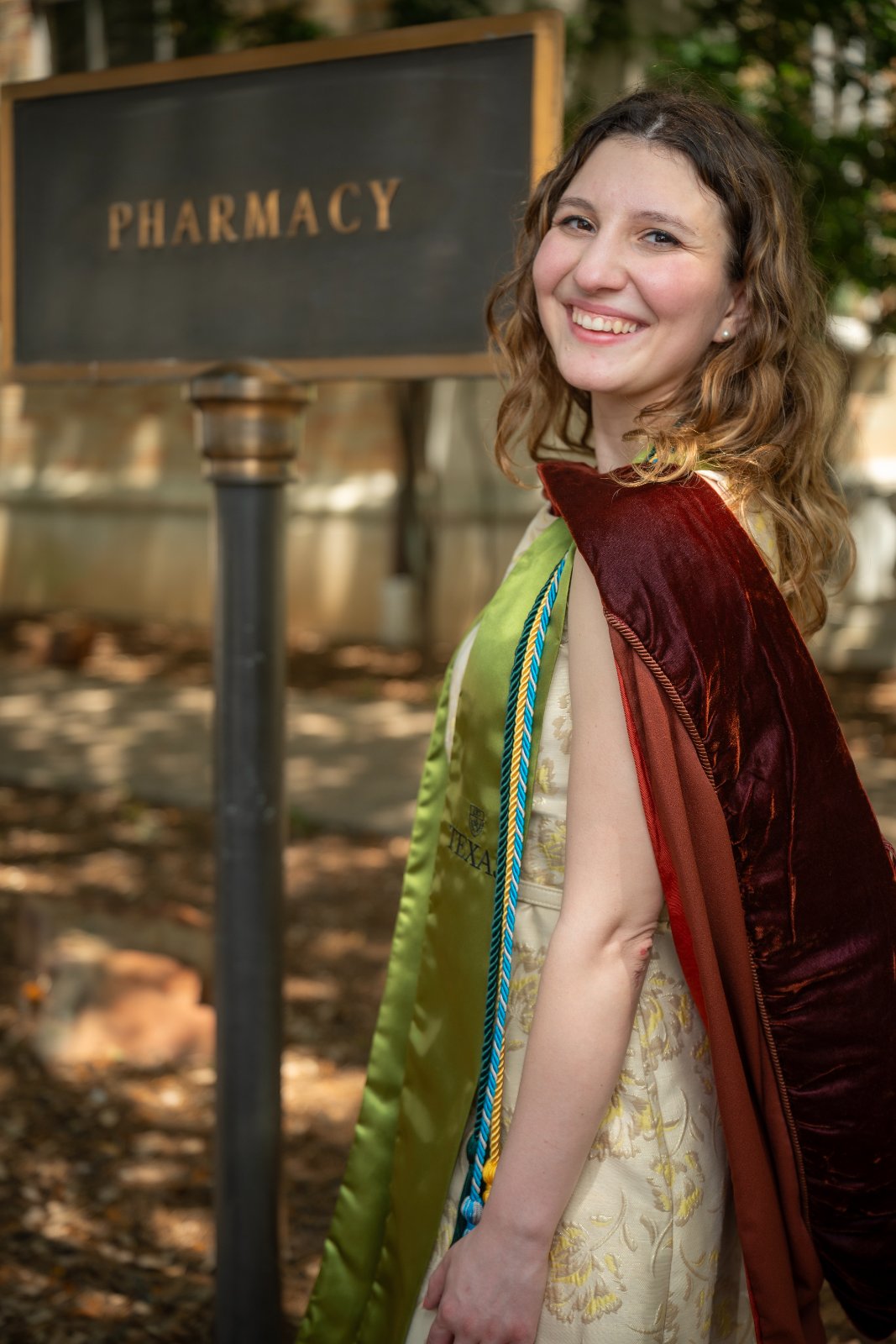 UT Austin pharmacy graduation portrait of the graduate in regalia standing beside the Pharmacy sign — photographed by Zan Visuals.