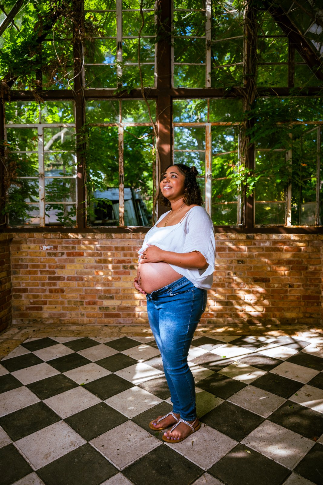 Maternity portrait of a pregnant woman standing in a sunlit indoor space with large windows and natural greenery in the background, gently holding her baby bump — photographed by Zan Visuals.