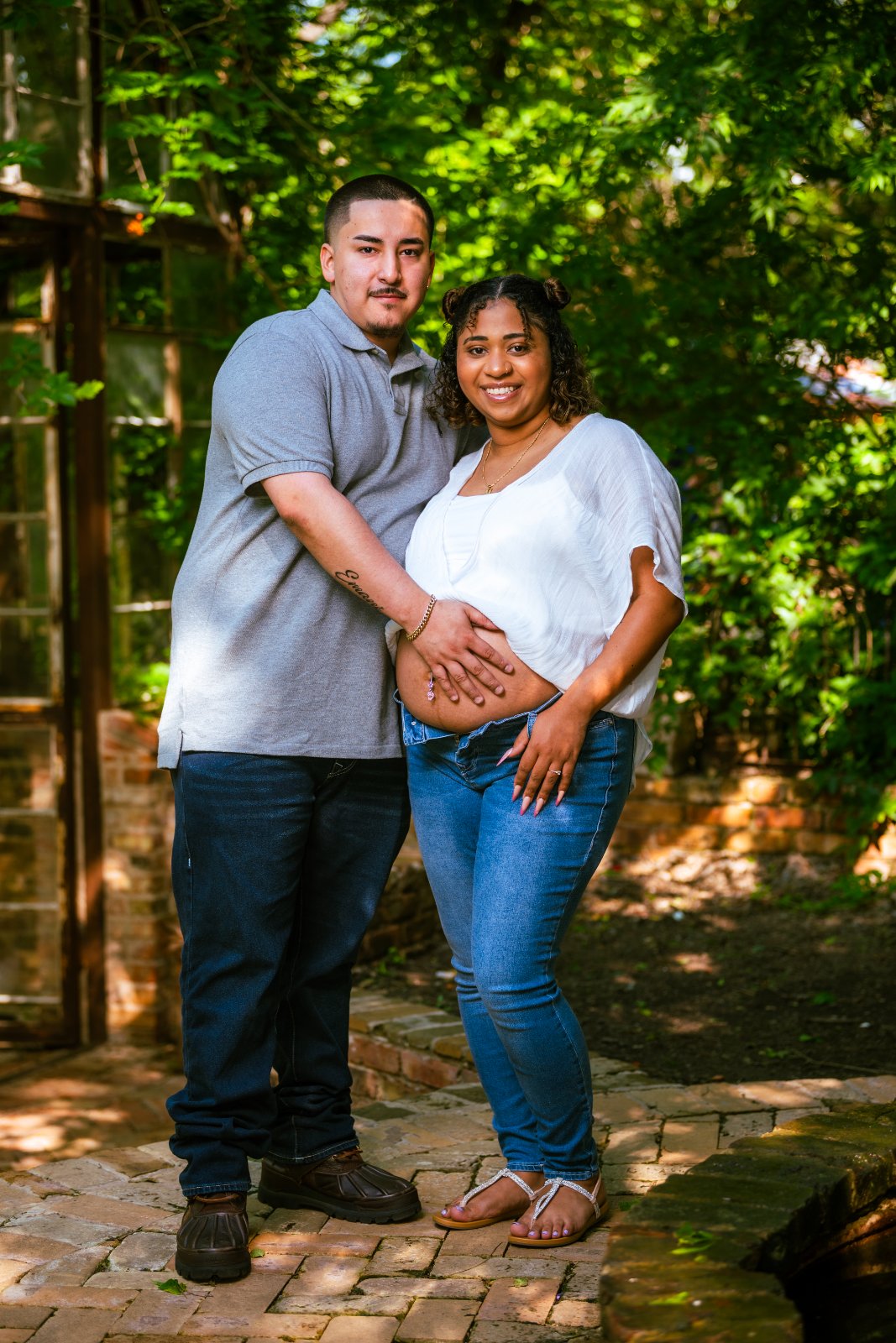 Couple holding the baby bump during a maternity photoshoot on a garden pathway surrounded by greenery — photographed by Zan Visuals.