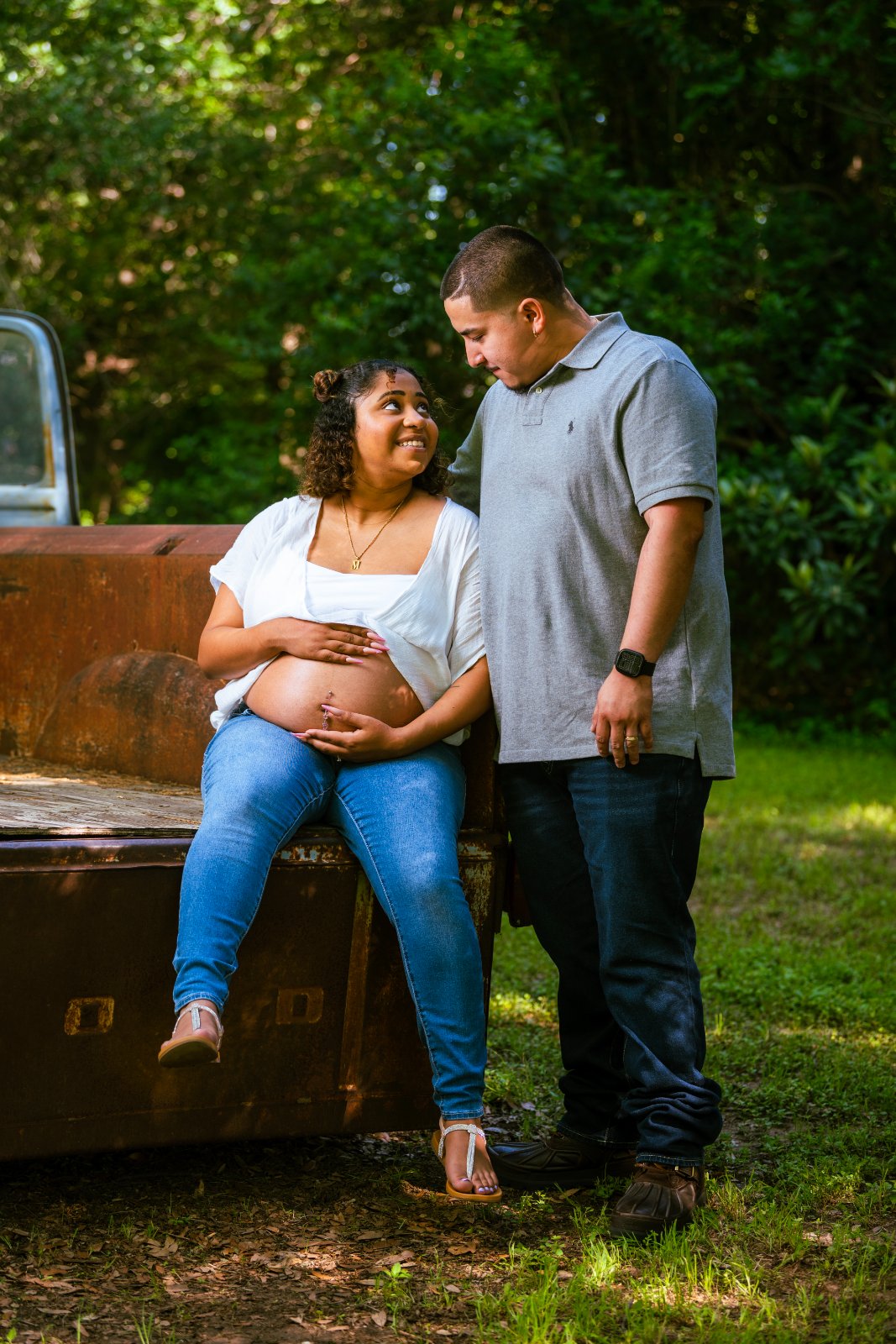 Couple sharing a quiet maternity moment beside an old truck, with hands resting on the baby bump in a natural outdoor setting — photographed by Zan Visuals.