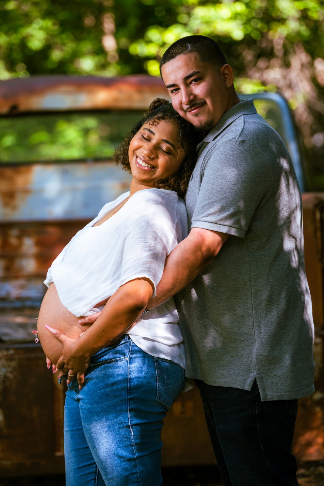 Couple embracing during a maternity photoshoot in front of a rustic vehicle, with hands gently resting on the baby bump in a warm outdoor setting — photographed by Zan Visuals.