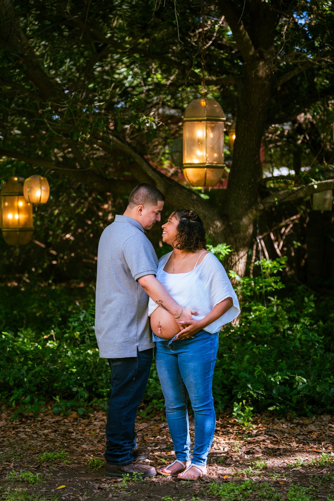 Couple holding the baby bump under hanging lanterns during a maternity photoshoot in a warm, nature‑filled outdoor setting — photographed by Zan Visuals.